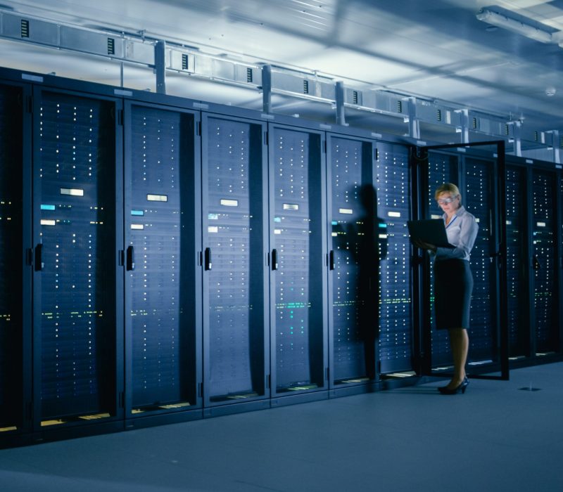 In Data Center: Female IT Technician Stands before Open Server Rack Cabinet, Uses Laptop Computer to Run Maintenance Diagnostics so that Mainframe Works at Optimal Functioning Level.